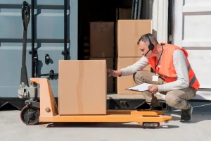 Warehouse worker inspecting shipment box during freight transport Canada operations at a loading dock with pallet truck and cargo container